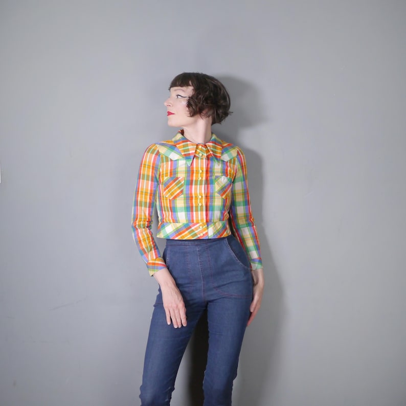 May include: A vintage, long-sleeved plaid shirt in a vibrant mix of yellow, orange, green, and blue. The shirt features a pointed collar and two chest pockets. The model is wearing the shirt with high-waisted blue jeans.