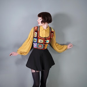 May include: A multi-coloured crochet vest with a square pattern, worn over a mustard yellow collared shirt and a black skirt. The vest features red, blue, green, and yellow squares. The outfit is completed with black stockings.