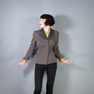 Puede incluir: Una blazer gris con cuello con cuentas y detalles de botones. La blazer se combina con una camisa amarilla y pantalones negros. Las mangas tienen puños de encaje amarillos. La blazer es de estilo vintage.