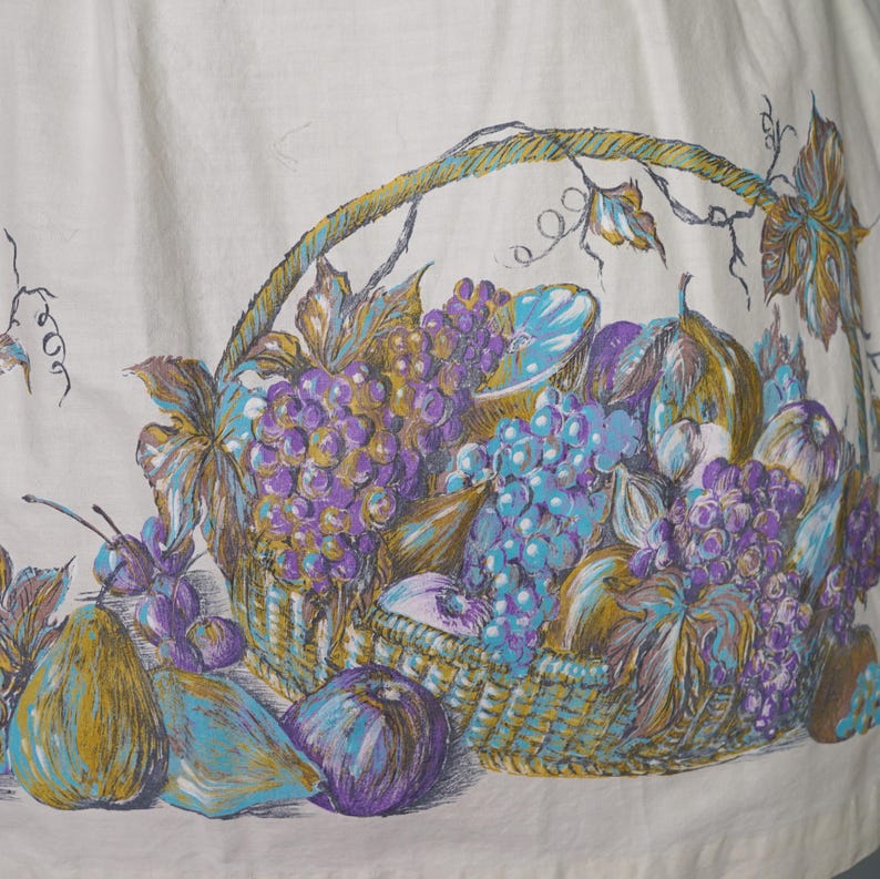 May include: A white fabric with a printed design of a basket overflowing with purple and blue grapes, pears, and other fruits. The basket is woven with brown and gold strands.