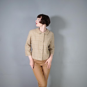 May include: A tan plaid jacket with a rounded collar and three matching buttons. The jacket has three-quarter sleeves and is paired with tan trousers. The outfit is set against a grey background.
