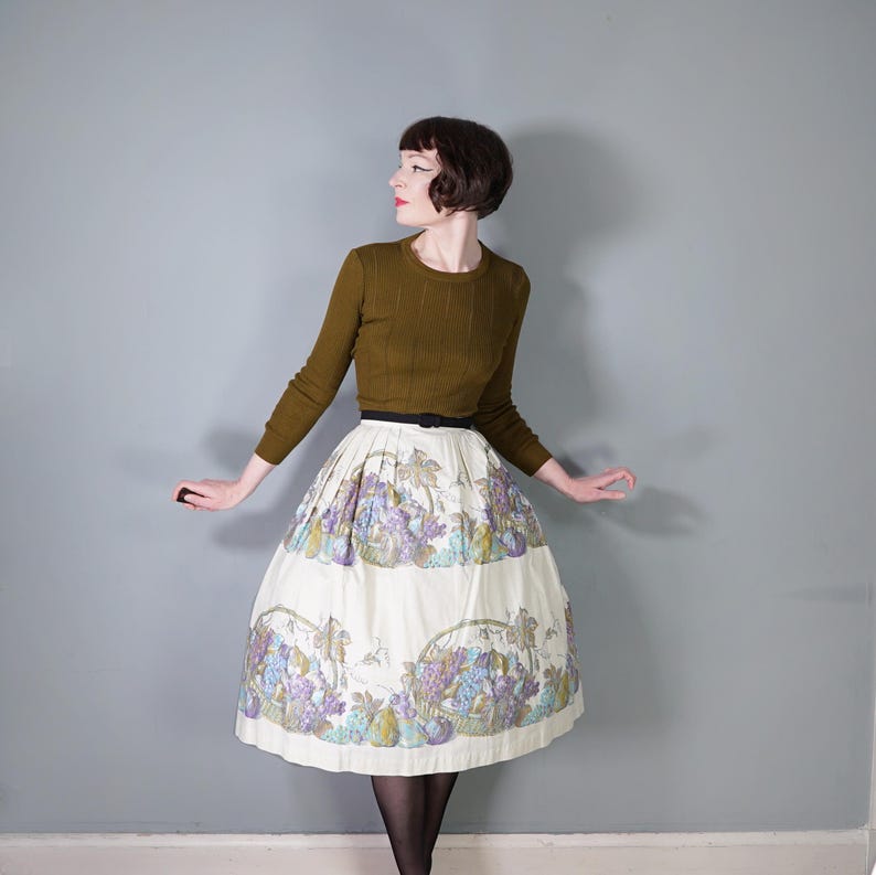 May include: A woman wearing a brown sweater and a white skirt with a floral print of grapes and pears. The skirt is full and has a black belt.