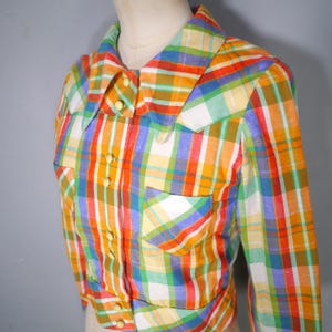 May include: A colorful plaid shirt with long sleeves, a collar, and two front pockets. The shirt features a vibrant pattern of red, orange, yellow, green, blue, and white stripes. It has small, round, yellow buttons down the front and on the cuffs.