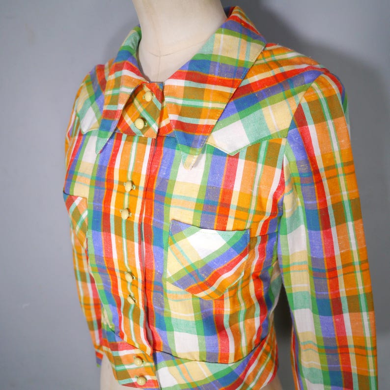 May include: A colorful plaid shirt with long sleeves, a collar, and two front pockets. The shirt features a vibrant pattern of red, orange, yellow, green, blue, and white stripes. It has small, round, yellow buttons down the front and on the cuffs.