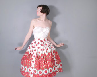 May include: A vintage strapless dress with a white bodice and a full skirt. The skirt features a white background with a repeating pattern of red flowers and green accents. The dress is knee-length and has a classic, retro style.