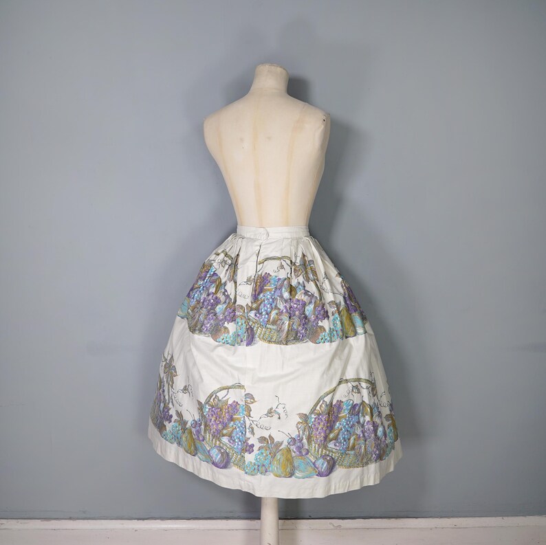 May include: A white skirt with a full gathered skirt silhouette. The skirt has a colorful print of fruit and grapes.