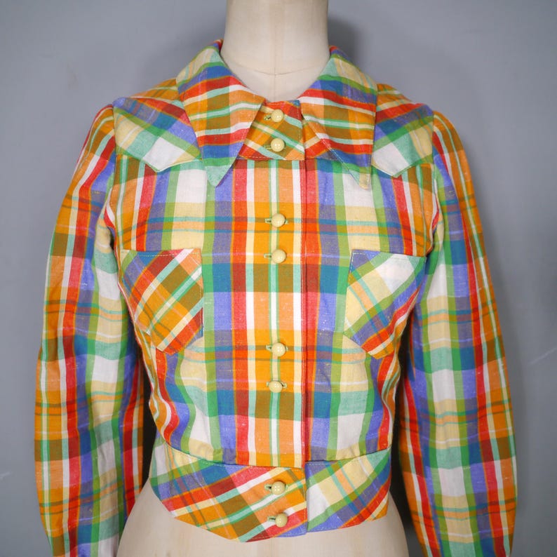 May include: A vintage, multi-colored plaid shirt with long sleeves, a collar, and two front pockets. The shirt features a button-up front and is displayed on a mannequin. The plaid pattern includes orange, yellow, green, blue, and white.
