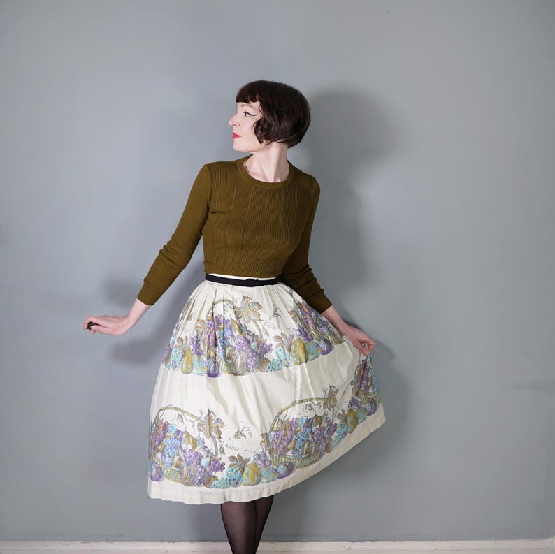 May include: A woman wearing a brown sweater and a white skirt with a floral print of grapes and pears. The skirt has a black belt.