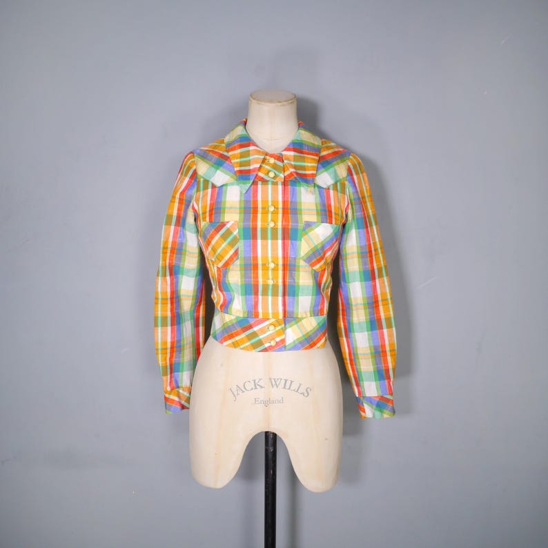 May include: A vintage, cropped plaid shirt in a rainbow of colors, including red, yellow, green, and blue. The shirt has long sleeves, a collared neckline, and two front pockets. The shirt is displayed on a mannequin.
