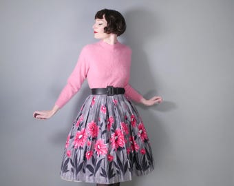 May include: A woman wearing a pink sweater and a gray skirt with pink flowers. She is wearing a black belt and has her hair styled in a short, curly bob.