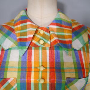 May include: A colorful plaid shirt in shades of orange, yellow, green, blue, and white. The shirt features a collared neckline, three cream-colored buttons, and a pocket. The fabric has a textured appearance.