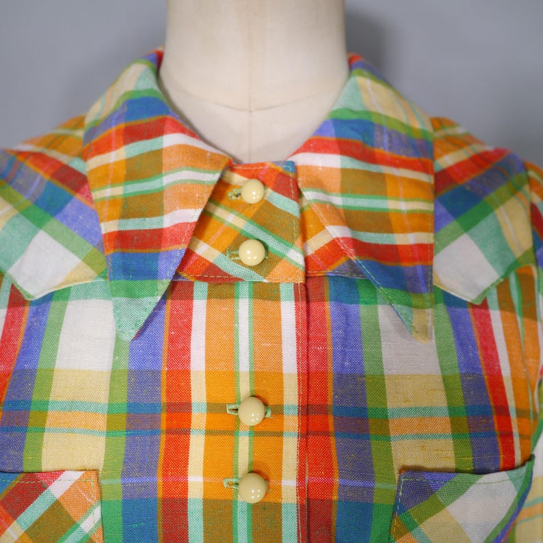 May include: A colorful plaid shirt in shades of orange, yellow, green, blue, and white. The shirt features a collared neckline, three cream-colored buttons, and a pocket. The fabric has a textured appearance.