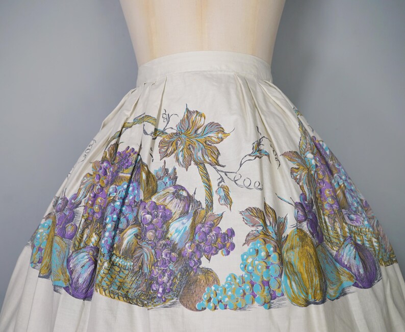 May include: A white full skirt with a vintage floral print featuring grapes, leaves, and fruit. The print is in shades of purple, blue, green, and brown.