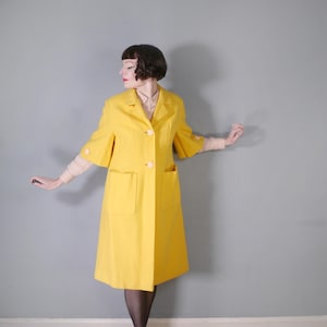 May include: A vibrant yellow coat featuring large front pockets and three buttons. The coat has short sleeves with button detailing and a classic collar. The knee-length coat appears to be made from a lightweight fabric.