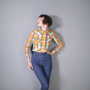 May include: A colorful plaid shirt with long sleeves and a collar, featuring a mix of yellow, orange, green, and red hues. The shirt is paired with high-waisted blue jeans.