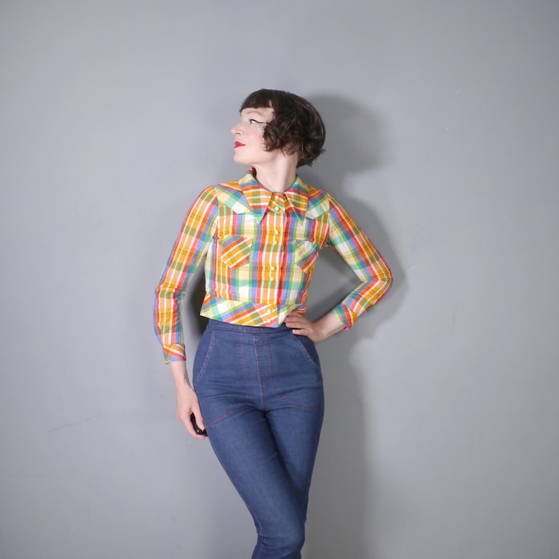 May include: A colorful plaid shirt with long sleeves and a collar, featuring a mix of yellow, orange, green, and red hues. The shirt is paired with high-waisted blue jeans.