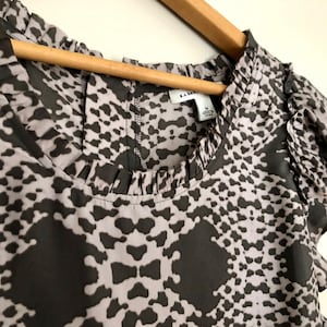 Puede incluir: Una blusa de estampado gris y negro con cuello de volantes y mangas cortas. La blusa tiene una etiqueta de talla que dice "Banana Republic" y "M".