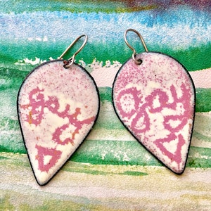 Puede incluir: Un par de pendientes en forma de lágrima con un diseño moteado blanco y rosa. Los pendientes tienen un borde negro y ganchos plateados. La palabra "JOY" es visible en rosa estilizado en cada pendiente.