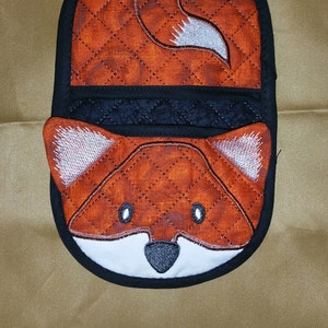 May include: A quilted orange and white fox pot holder with a black border. The fox's face is visible with white ears, black eyes, and a black nose.
