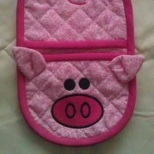 Pode incluir: Luva de forno acolchoada rosa com um design de rosto de porco. O porco tem olhos pretos, um focinho rosa com duas narinas e orelhas rosas.