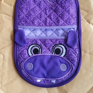 May include: Purple quilted pot holder with a stitched design of a hippopotamus face. The hippo has large eyes, a wide mouth, and a small nose.