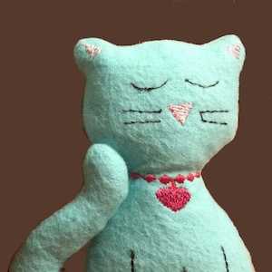 May include: A light blue plush cat toy with embroidered eyes, a pink nose, and a red heart on a necklace. The cat is sitting with its paw raised.