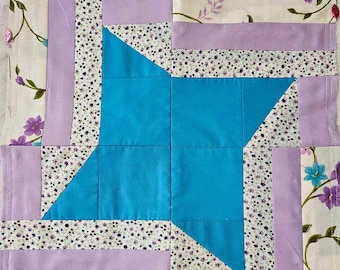 May include: A quilt block featuring a blue star design, surrounded by a border of purple and white fabric with a floral pattern.