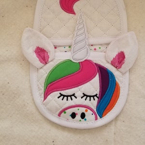 May include: White quilted pot holder with a unicorn design. The unicorn has a pink mane, a green horn, and a rainbow tail. The unicorn's eyes are closed and it has a black nose.