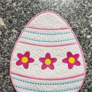 Può includere: Un sottopentola bianco trapuntato a forma di uovo di Pasqua con ricami rosa e blu. L'uovo ha tre fiori rosa con centri gialli.