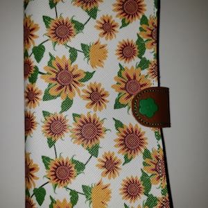 以下が含まれることがあります： ひまわり柄の白い財布。茶色のフラップに緑の花のボタンが付いている。