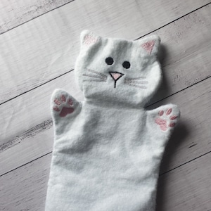 May include: White felt hand puppet with a cat face. The cat has pink ears, a black nose, and gray whiskers. The puppet has pink paw prints on the hands.
