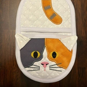 Pode incluir: Pegador de panela acolchoado branco com um rosto de gato calico e uma cauda nas costas. O gato tem pêlo laranja, cinza e branco com olhos amarelos e um nariz rosa.