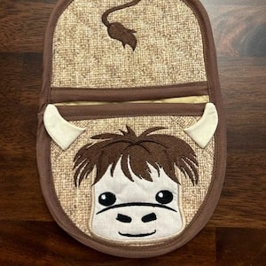 May include: Brown and white pot holder with a cow's face embroidered on it. The cow has a brown nose, brown eyes, and a brown tail.