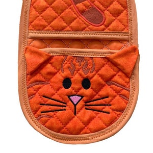 Pode incluir: Luva de forno acolchoada laranja com um design de rosto e cauda de gato. O rosto do gato tem olhos pretos, um nariz rosa e bigodes brancos.