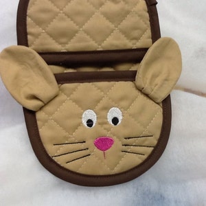May include: A brown quilted oven mitt with a cute embroidered cat face. The cat has white eyes, a pink nose, and brown whiskers.