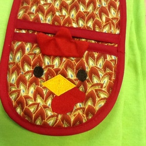 May include: Red and gold fabric pot holder with a rooster design. The rooster has a red comb, yellow beak, and black eyes. The pot holder is shaped like a rooster's head.