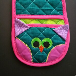 May include: A pink and green quilted hot pad with an owl design. The owl has purple ears, green eyes, and an orange beak.
