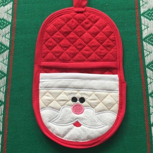 May include: Red quilted oven mitt with a white Santa Claus face embroidered on the front. The Santa has a white beard, black eyes, and a pink nose.