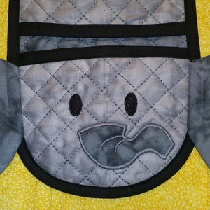 May include: A grey quilted oven mitt with a black trim and an embroidered elephant face. The elephant has large ears, black eyes, and a grey trunk.