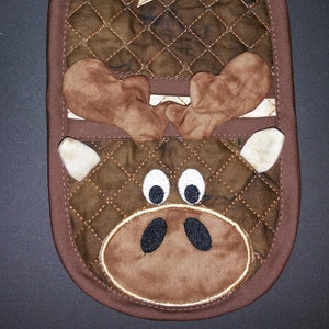 Pode incluir: Pegador de panela acolchoado marrom com um design de alce. O alce tem pelo marrom, chifres brancos e olhos pretos. O pegador de panela tem um laço marrom para pendurar.