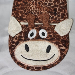 May include: A brown and tan pot holder shaped like a giraffe's face. The pot holder has a smiling mouth, two large eyes, and two ears.