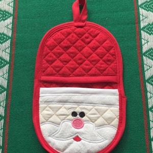 Puede incluir: Manopla de horno acolchada roja con una cara de Papá Noel blanca bordada en la parte delantera. Papá Noel tiene barba blanca, ojos negros y nariz rosa.
