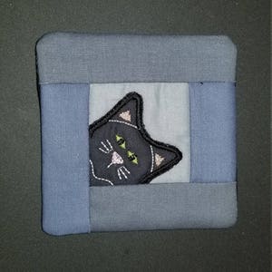 Pode incluir: Um quadrado de tecido azul com um rosto de gato preto bordado no centro. O gato tem olhos verdes e um nariz rosa.