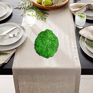 May include: A green leaf-shaped coaster with a detailed design. The coaster is on a white linen table runner.