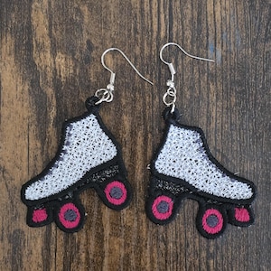 May include: A pair of silver roller skate earrings with white, black, and pink details. The skates are embroidered with a white and black pattern and have pink wheels.