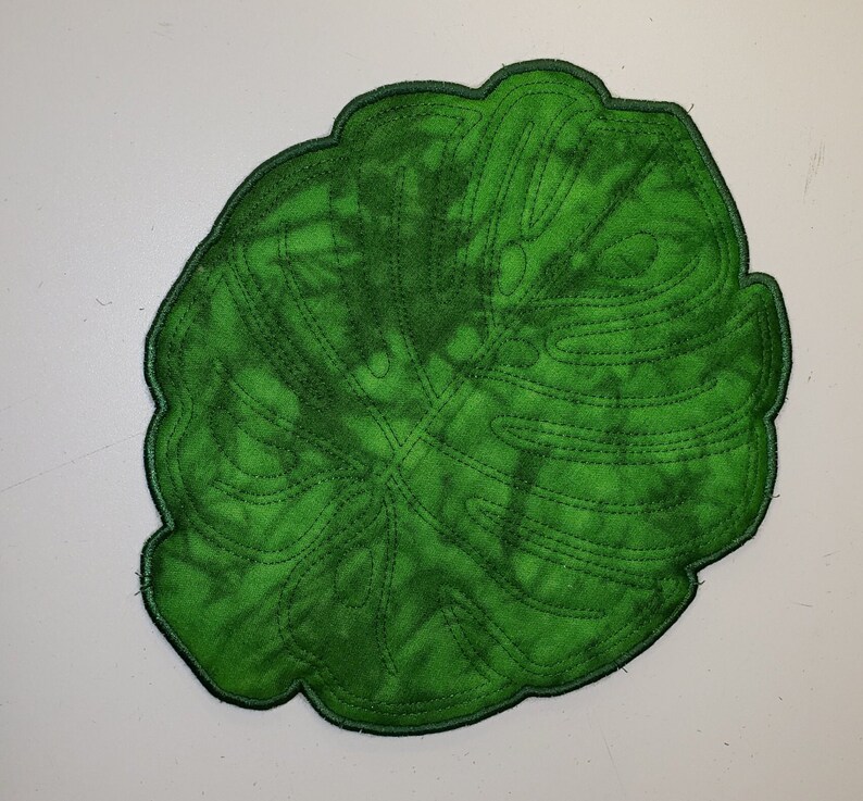 May include: A green felt monstera leaf with a stitched outline. The leaf is a light green colour with a darker green outline.