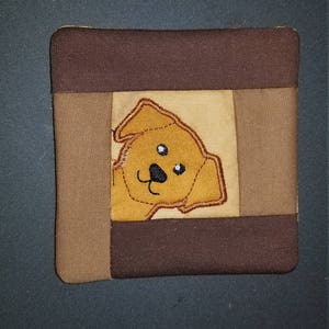 May include: A brown and tan fabric coaster with a brown embroidered dog peeking out from the centre.