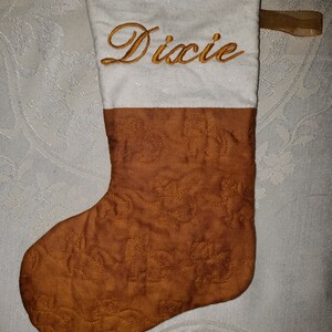 May include: A white and brown quilted Christmas stocking with the name "Dixie" embroidered in gold thread.