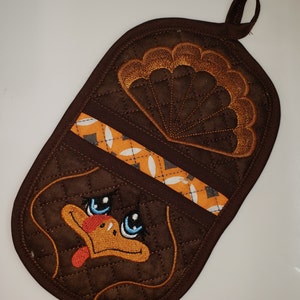 Pode incluir: Luva de forno acolchoada marrom com um rosto de peru e penas de cauda bordadas. O peru tem olhos azuis e um bico laranja.