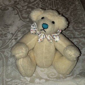 May include: A white plush teddy bear with a colorful polka dot bow around its neck. The bear has black eyes and a blue nose.